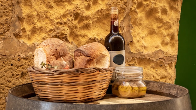 Bread in a basket next to a wine bottle and a jar of cheese on a wooden barrel