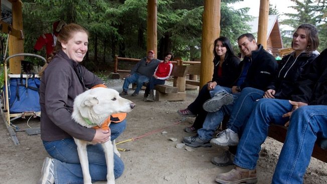 Um guia acariciando um cachorro puxador de trenó enquanto faz uma apresentação para um grupo de visitantes Um guia acariciando um cachorro puxador de trenó enquanto faz uma apresentação para um grupo de visitantes