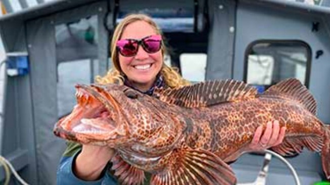 A smiling woman holding a lingcod fish on a fishing boat