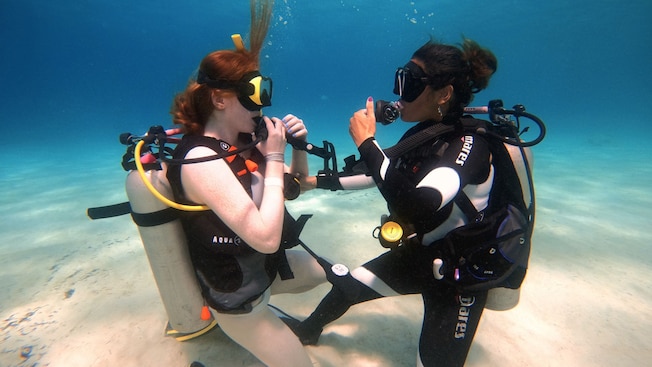 Two women in scuba diving gear kneeling on a sandy ocean floor Two women in scuba diving gear kneeling on a sandy ocean floor