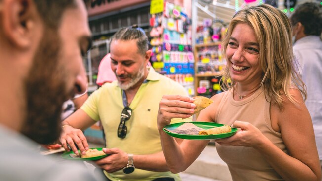 A smiling woman holding a plate of tacos while 2 other Guests enjoy their food at an indoor market