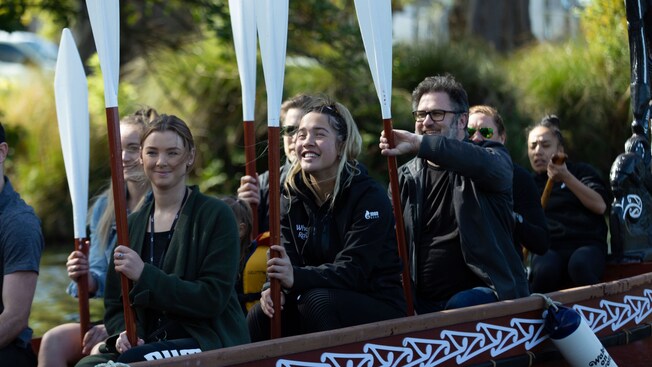 Visitantes sonrientes sentados en una tradicional waka maorí sosteniendo remos en un río