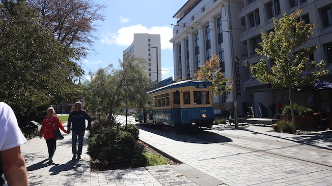 The Christchurch Tram operating on the city tour route with pedestrians, trees and buildings on both sides The Christchurch Tram operating on the city tour route with pedestrians, trees and buildings on both sides