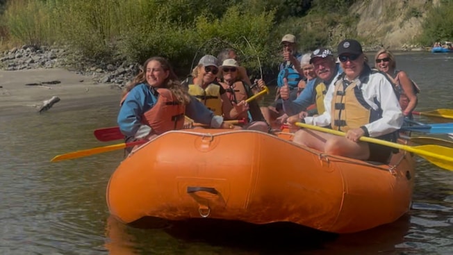 Group of smiling Guests in life jackets raft on a river near a sandy shore and forested hills Group of smiling Guests in life jackets raft on a river near a sandy shore and forested hills