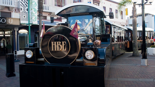 A vintage road train with the letters ‘H B E’ parked on a brick street surrounded by shops A vintage road train with the letters ‘H B E’ parked on a brick street surrounded by shops