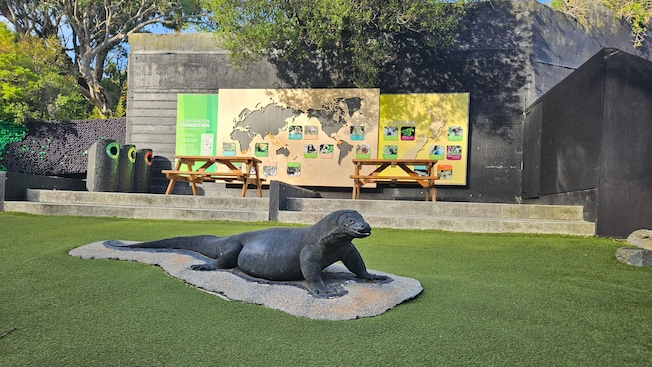 A Komodo dragon statue on the grass near a wall with a world map, animal pictures and 2 picnic tables A Komodo dragon statue on the grass near a wall with a world map, animal pictures and 2 picnic tables