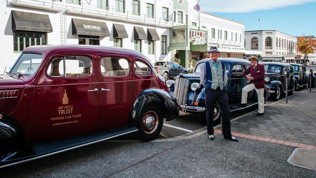 Vintage cars from Art Deco Trust, Vintage Car Tours, lined up on a street with drivers in period clothing