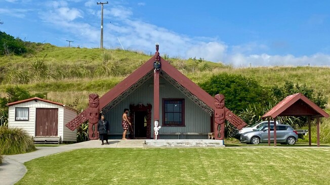 Two Māori people standing in front of a traditional gathering hall with wood carvings