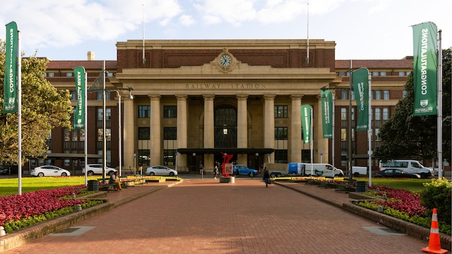 A brick pathway leading to a large neoclassical building with tall columns and a central clock and the words ‘Railway Station’ A brick pathway leading to a large neoclassical building with tall columns and a central clock and the words ‘Railway Station’