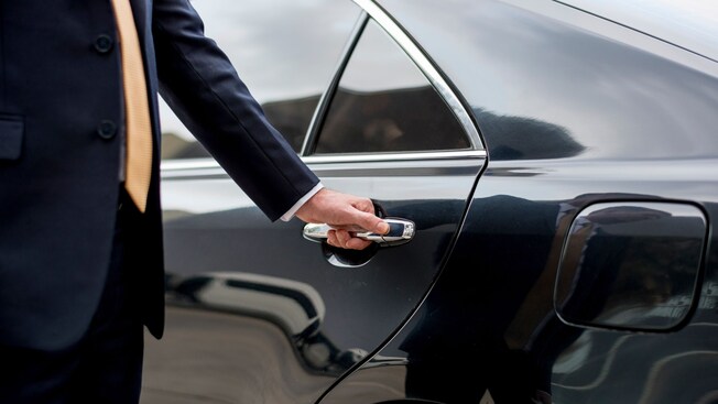 A professional driver in a suit opening the rear door of a luxury sedan