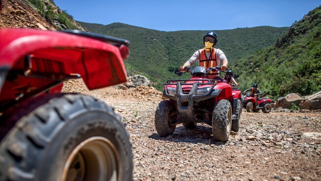 A rider wearing a helmet, goggles and face mask driving in a caravan of all terrain vehicles over mountainous terrain