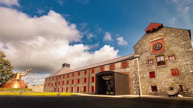 Uma grande chaleira de cobre em frente a um antigo edifício de tijolos e as palavras, "Midleton Distillery Experience" Uma grande chaleira de cobre em frente a um antigo edifício de tijolos e as palavras, "Midleton Distillery Experience"