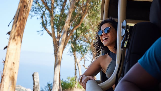 A smiling woman leaning out of a safari style vehicle on a road with trees and the ocean in the background A smiling woman leaning out of a safari style vehicle on a road with trees and the ocean in the background
