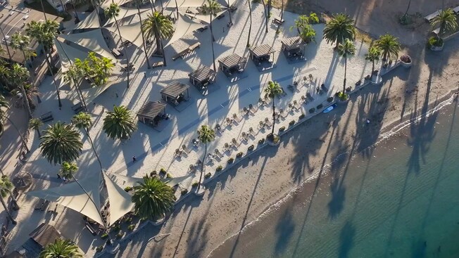 An aerial view of a private beach with palapas, picnic tables, palm trees and lounge chairs