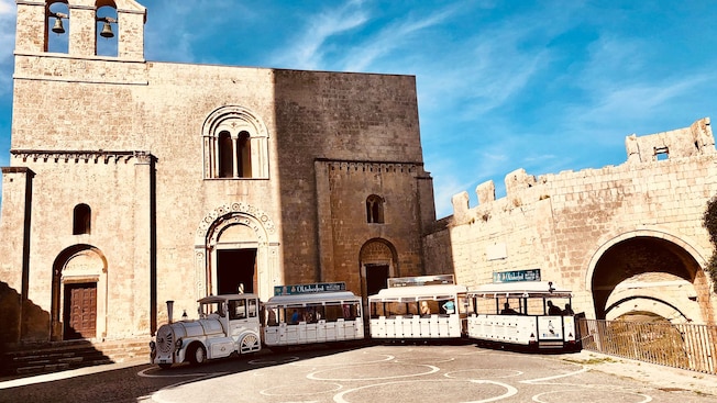 Edificio histórico de piedra con ventanas arqueadas y campanarios, y un autobús turístico estacionado frente a él.