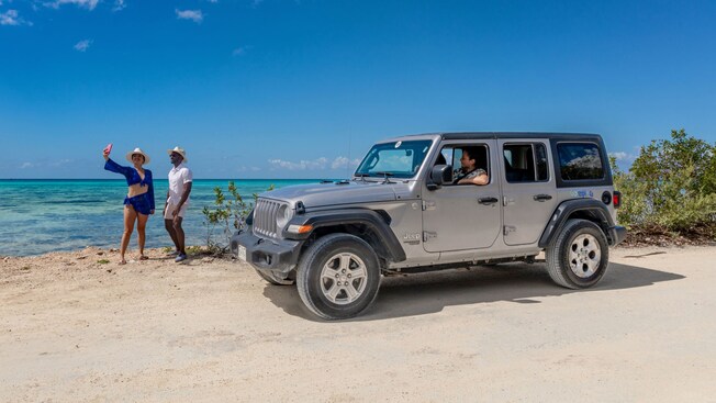 Two people taking a selfie on a beach near a parked off road vehicle with a woman in the driver seat