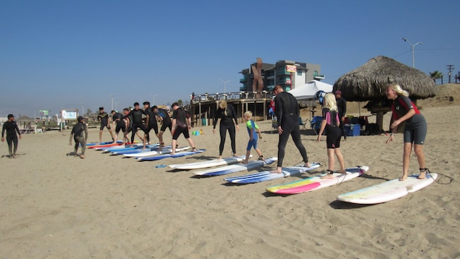 Un grupo de surfistas en trajes de neopreno de pie sobre sus tablas en la playa, con un instructor guiándolos Un grupo de surfistas en trajes de neopreno de pie sobre sus tablas en la playa, con un instructor guiándolos