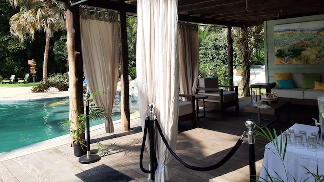 A cabana with a sofa, snacks on a coffee table, a cucumber infused water station and padded chairs next to a pool A cabana with a sofa, snacks on a coffee table, a cucumber infused water station and padded chairs next to a pool