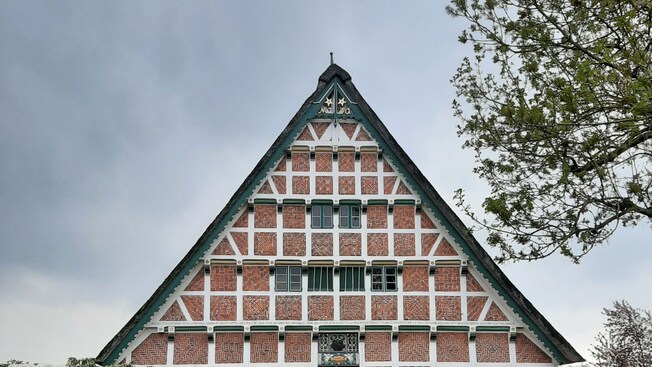 An A shaped house with an edifice featuring wood that separates squares of patterned bricks, an open front door, blooming flowers and a manicured lawn