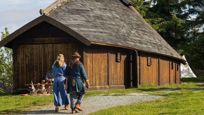 A man and a woman dressed in traditional Viking clothing, walking towards a traditional Viking farmhouse