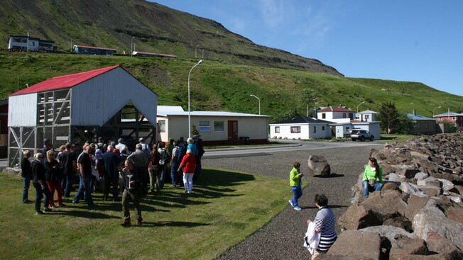 People gathered outside a silo like building between a hillside and a jetty