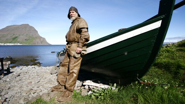 Un pescador vestido con ropa artesanal y mocasines se apoya en un bote de remos