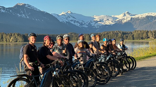 14 smiling people standing beside their electronic bikes at the edge of a lake on North Douglas Island in Alaska