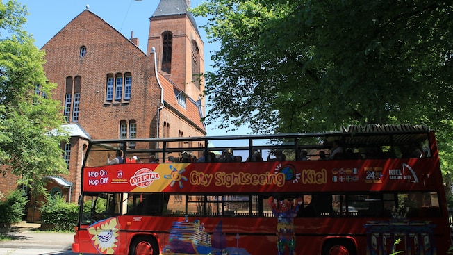 A double decker tour bus with the words ‘City Sightseeing Kiel’ driving past a brick cathedral A double decker tour bus with the words ‘City Sightseeing Kiel’ driving past a brick cathedral