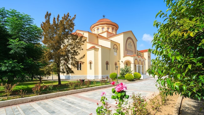 Un monasterio soleado con un camino pavimentado rodeado de flores, árboles y podas ornamentales Un monasterio soleado con un camino pavimentado rodeado de flores, árboles y podas ornamentales