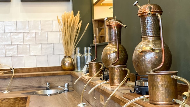 Una estación de bar con fregadero incorporado y tres destiladores de ginebra de cobre alineados contra la pared Una estación de bar con fregadero incorporado y tres destiladores de ginebra de cobre alineados contra la pared