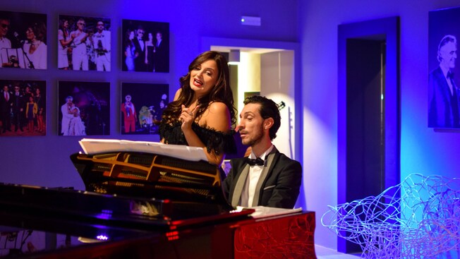 Performer in an elegant gown singing beside a pianist in formal attire, with portraits and vibrant lighting