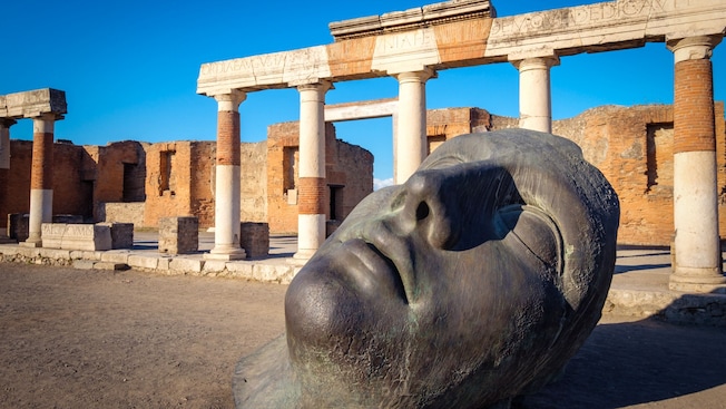 Cabeça grande de uma estátua deitada no chão em frente a estruturas históricas em Pompeia Cabeça grande de uma estátua deitada no chão em frente a estruturas históricas em Pompeia