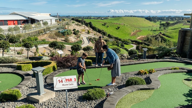 Dos mujeres y un niño jugando minigolf en la cima de un campo montañoso y un letrero que dice: ‘Par 3’ Dos mujeres y un niño jugando minigolf en la cima de un campo montañoso y un letrero que dice: ‘Par 3’