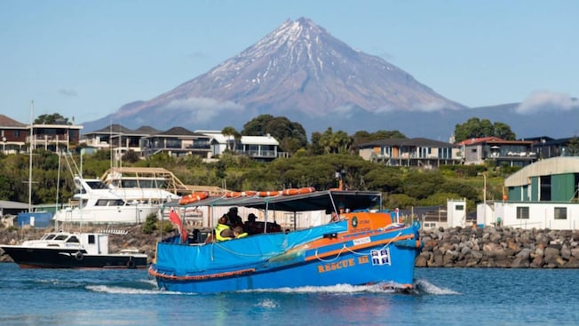 A tour boat setting off in a port in front of oceanside houses and Mount Taranaki A tour boat setting off in a port in front of oceanside houses and Mount Taranaki