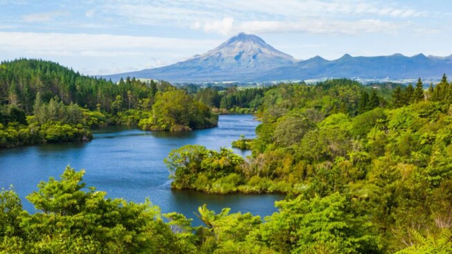An aerial view of 3 people waving in a lush landscape with a lake and a mountain in the distance