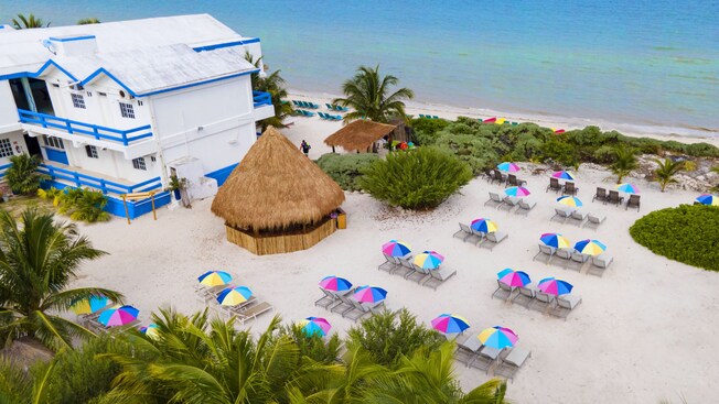 A modern 2 story building, a few thatched roof buildings and an array of recliners on a sandy beach