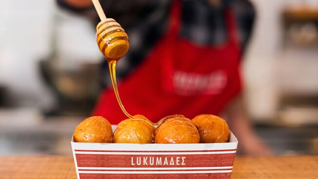 A food box of fritters with honey drizzling down labeled 'Loukoumades Authentic Greek Delights'