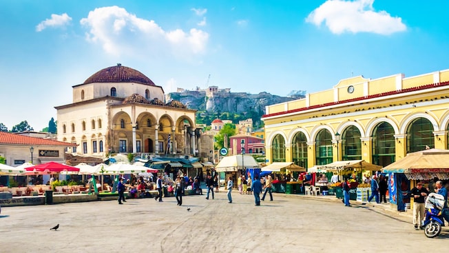 A group of people walking in a town square with buildings and a dome in Monastiraki in Athens, Greece A group of people walking in a town square with buildings and a dome in Monastiraki in Athens, Greece