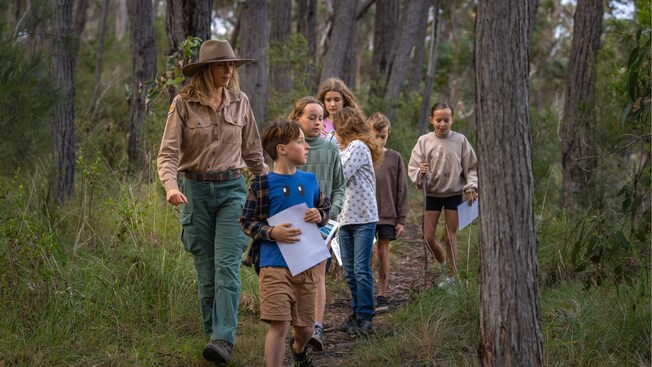 A National Parks Guide leading a group of children down a trail through some trees