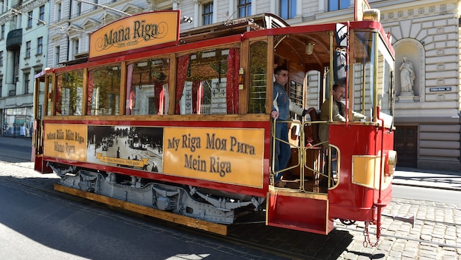 A trolley car with a sign that reads Mana Riga stopped next to a neoclassical building A trolley car with a sign that reads Mana Riga stopped next to a neoclassical building