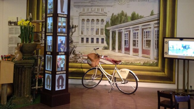 A museum display featuring a bicyle, a tower clock, a video display and a large mural of 2 neoclassical buildings