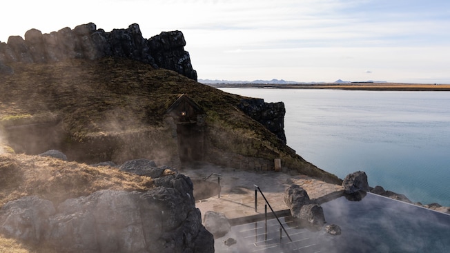 Uma entrada construída em um afloramento rochoso, um caminho de pedra e escadas que descem para uma lagoa fumegante Uma entrada construída em um afloramento rochoso, um caminho de pedra e escadas que descem para uma lagoa fumegante