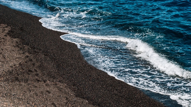 Olas suaves chocando contra una playa de arena negra en la isla de lava de Nea Kameni, situada en el corazón de la caldera de Santorini Olas suaves chocando contra una playa de arena negra en la isla de lava de Nea Kameni, situada en el corazón de la caldera de Santorini