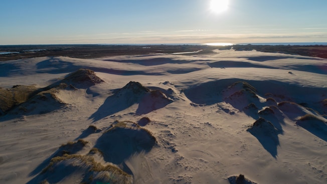 A sand dune stretches towards a large body of water on the horizon as the sun sets A sand dune stretches towards a large body of water on the horizon as the sun sets