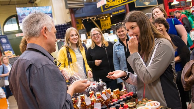 A Guest samples food while a shopkeeper and other Guests watch A Guest samples food while a shopkeeper and other Guests watch