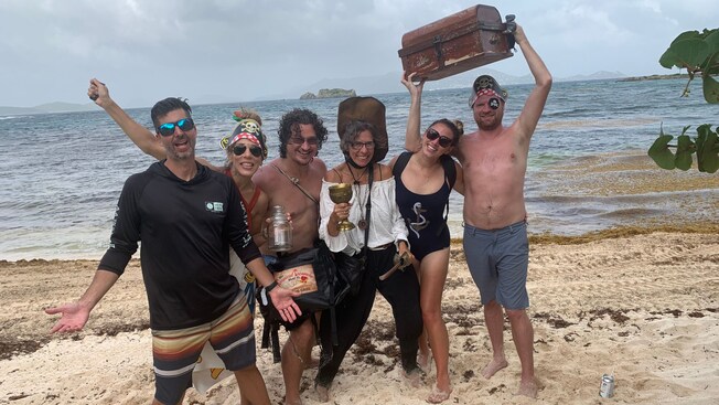Five Guests and an actor playing Anne Bonny standing on a beach with a treasure chest, sword and goblet