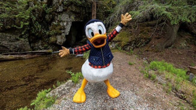 Donald Duck wearing a lumberjack outfit in a wide arm pose next to a creek in Alaska