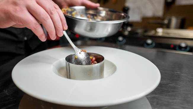 A chef plating food using a metal ring mold and spoon in a professional kitchen A chef plating food using a metal ring mold and spoon in a professional kitchen