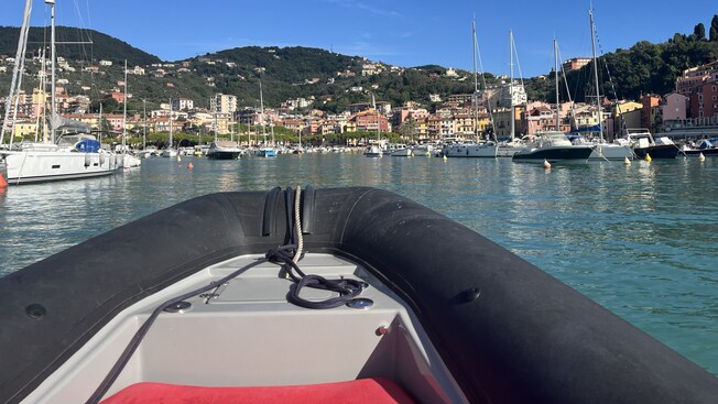 View from the bow of a rubber boat approaching a harbor with sailboats and buildings on a hillside