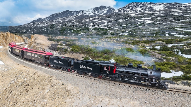 A steam train with passenger cars traveling along a mountain railway, with rugged peaks and patches of snow in the background A steam train with passenger cars traveling along a mountain railway, with rugged peaks and patches of snow in the background